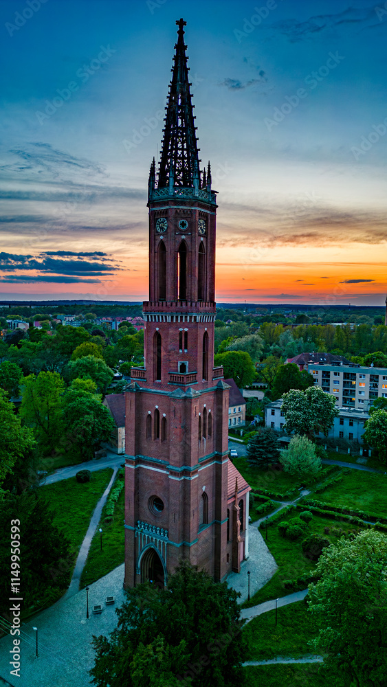 Fototapeta premium Sunset Over Gothic Church Tower in Żagań, Poland. Aerial view of a towering gothic-style church in Żagań, Poland, surrounded by greenery and urban scenery, set against a breathtaking sunset sky.