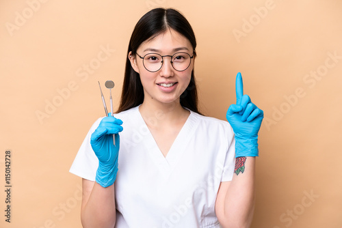 Dentist Chinese woman holding tools isolated on beige background pointing up a great idea