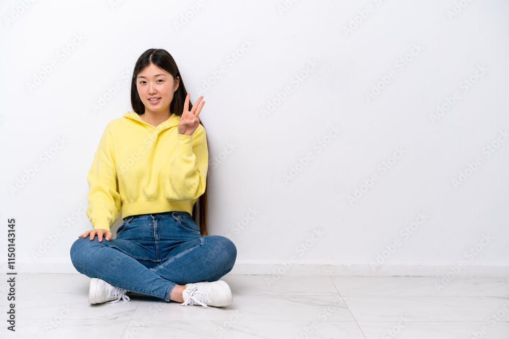 Young Chinese woman sitting on the floor isolated on white wall happy and counting three with fingers Young Chinese woman sitting on the floor isolated on white wall happy and counting three with fingers