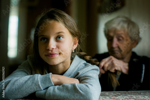 A grandmother braids her granddaughter's hair as the teenager sits with a slightly displeased expression, captured in a quiet, intimate moment.
