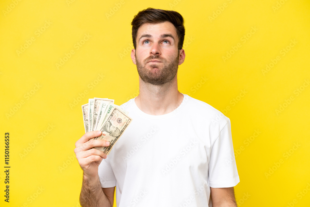 Young caucasian man taking a lot of money isolated on yellow background and looking up Young caucasian man taking a lot of money isolated on yellow background and looking up