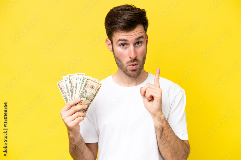 Young caucasian man taking a lot of money isolated on yellow background intending to realizes the solution while lifting a finger up Young caucasian man taking a lot of money isolated on yellow background intending to realizes the solution while lifting a finger up