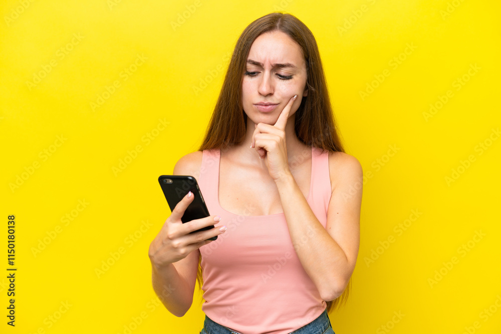 Young caucasian woman isolated on yellow background using mobile phone and thinking