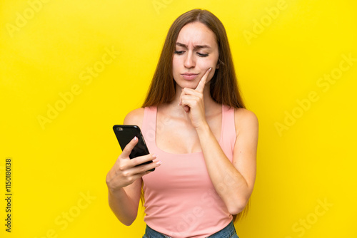 Young caucasian woman isolated on yellow background using mobile phone and thinking