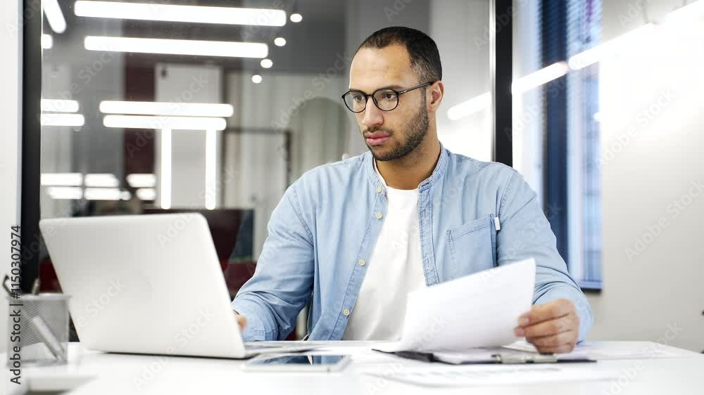 Busy thoughtful African American businessman analyzing financial documents checking data on laptop while sitting at a desk at a workplace in a business office. Financier deals with financial report