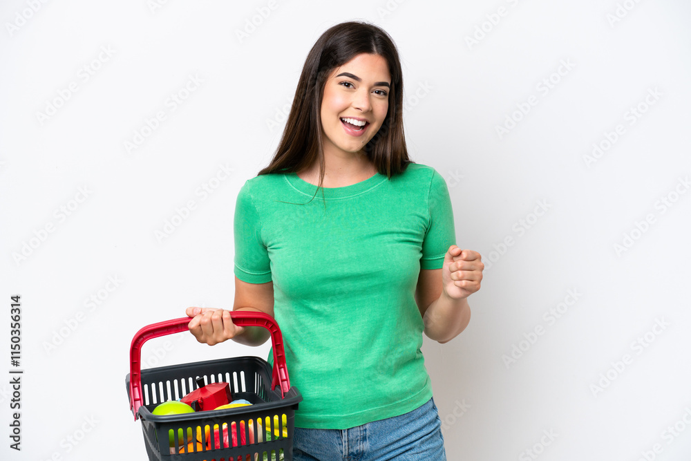 Young Brazilian woman holding a shopping basket full of food isolated on white background celebrating a victory in winner position Young Brazilian woman holding a shopping basket full of food isolated on white background celebrating a victory in winner position