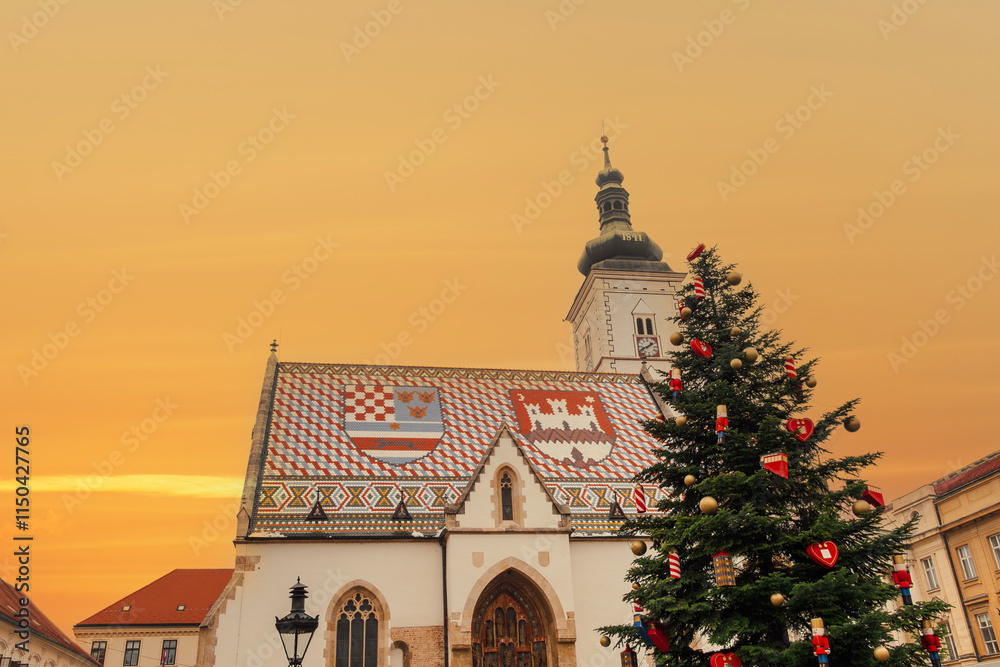 Fototapeta premium Zagreb, Croatia evening view of a beautifully adorned Christmas tree standing in front of St Mark's Church with its emblematic roof under a vivid orange sky at sunset.
