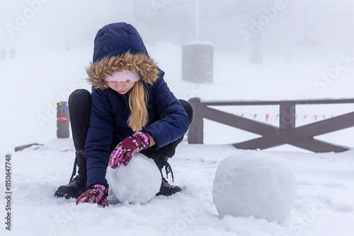Girl Building Snowman on Foggy Winter Day. A young girl in a winter coat and gloves rolls a snowball while building a snowman on a foggy snowy day.