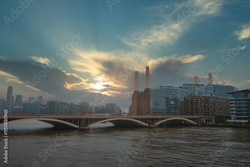 Battersea Power Station's chimneys rise against a dramatic sky as the sun sets, casting a warm glow over the River Thames and modern buildings. photo