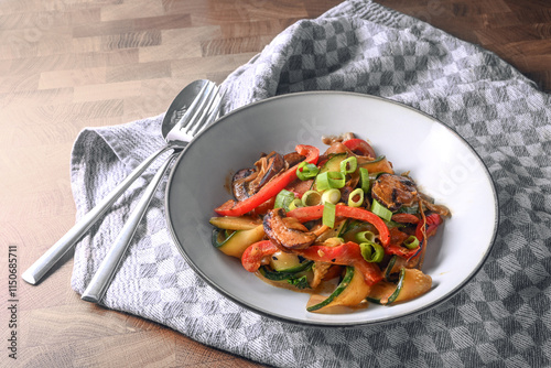 Vegetable dish with red bell pepper, zucchini, mushrooms and spring onion garnish in a plate with cutlery on a gray kitchen towel and a wooden table, copy space, selected focus