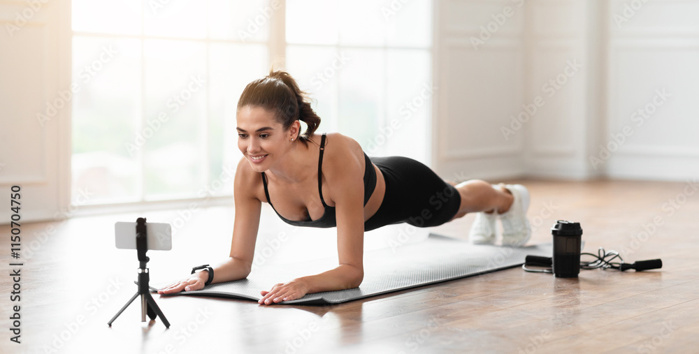 Fototapeta premium Happy young lady in sportswear planking in living room or fitness studio, watching video or having online conference with trainer, sporty woman exercising and looking at smartphone screen, free space