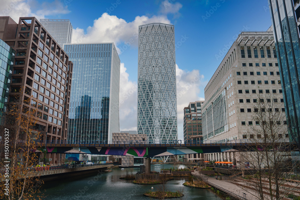 Naklejka premium View of Canary Wharf in London, featuring high rise buildings and a cylindrical skyscraper. A colorful bridge spans a waterway under a partly cloudy sky.