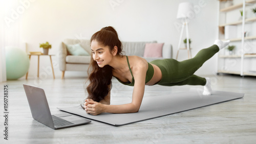Strong Indian woman standing in elbow plank, lifting her leg on sports mat, following online video tutorial on laptop at home. Young Eastern lady exercising her core muscles, doing domestic fitness