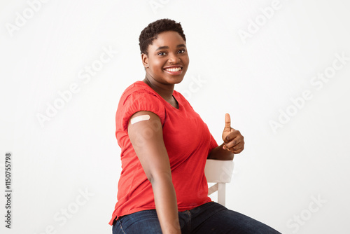 Coronavirus Vaccination. Happy African Lady Showing Vaccinated Arm With Adhesive Plaster After Antiviral Covid-19 Vaccine Shot, Gesturing Thumbs-Up Approving Vaccinating On White Studio Background photo