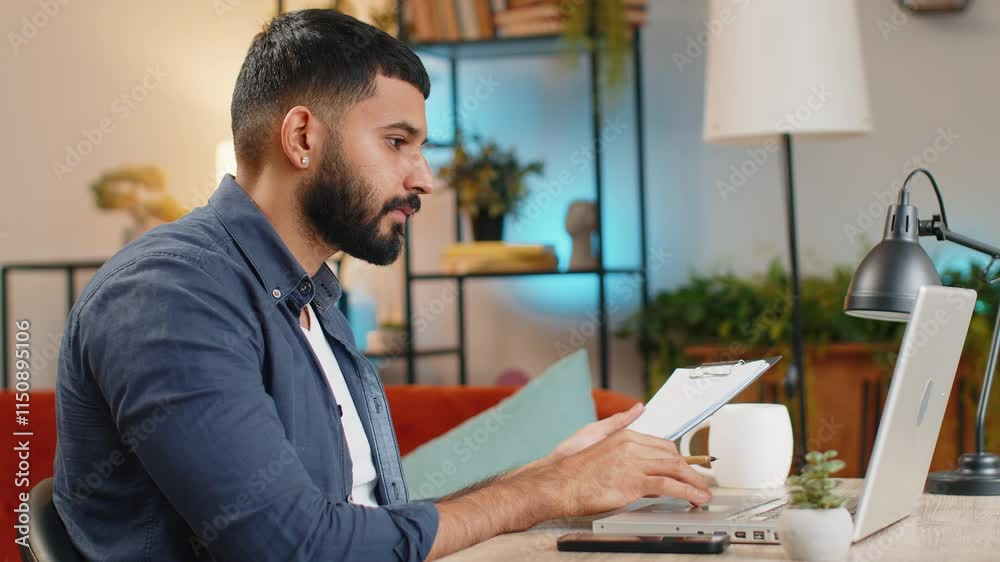 Indian young bearded freelancer making notes on clipboard at home office, using laptop, reading information prepare business financial report. Guy analyzing reports documents calculate taxes at table.