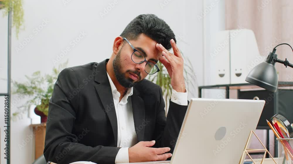 Tired ill Indian businessman suffering from headache problem tension and migraine, stress at home office desk workplace. Bearded freelancer broker worker man works on laptop notebook. Overworking