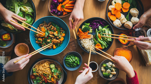 Overhead view of a group of people sharing a variety of colorful Asian dishes using chopsticks, symbolizing communal dining and culture.