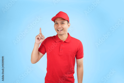 An enthusiastic Asian courier in a red uniform and cap points upwards with a bright smile, highlighting a service offer or promotional message in a studio setting with a blue background.