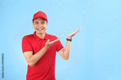 An enthusiastic Asian courier in a red uniform and cap points upwards with a bright smile, highlighting a service offer or promotional message in a studio setting with a blue background.