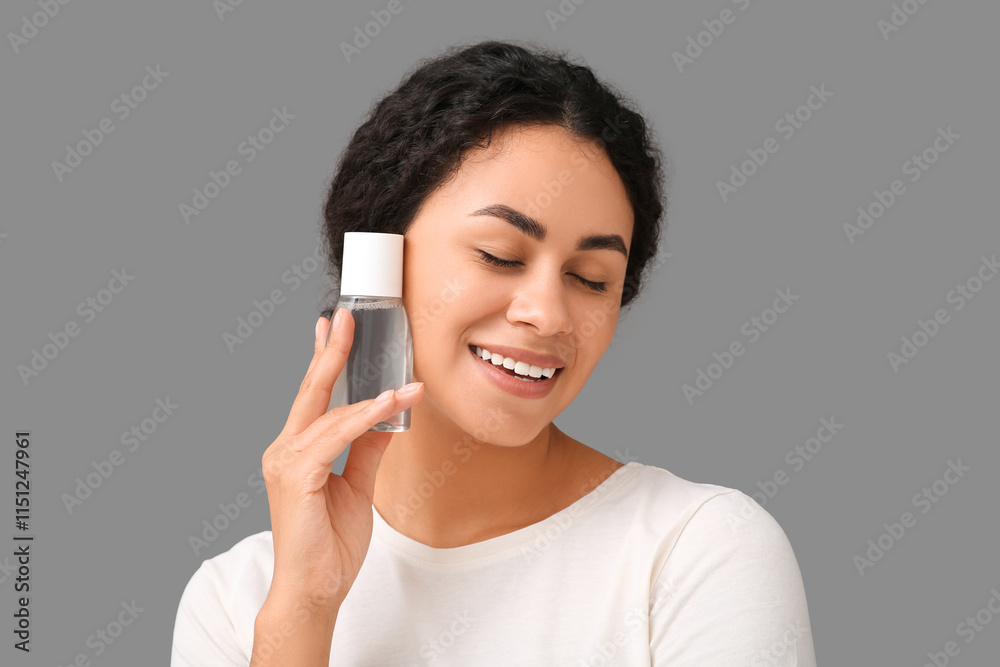 Happy young African-American woman with bottle of micellar water on grey background Happy young African-American woman with bottle of micellar water on grey background
