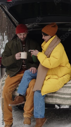 Vertical shot of couple cosily nesting in open car trunk, with hot thermos of tea, talking and enjoying relaxing wintry day