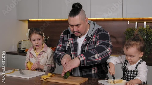 Caring handsome stylish father and two lovely elementary age daughter preparing fresh fruit salad, peeling banana and cutting tasty avocado on chopping boards while family cooking in kitchen.