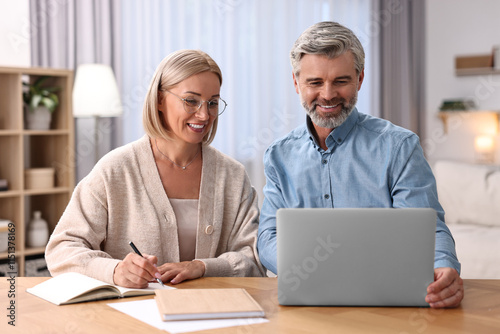 Happy middle aged couple working with laptop at wooden table indoors