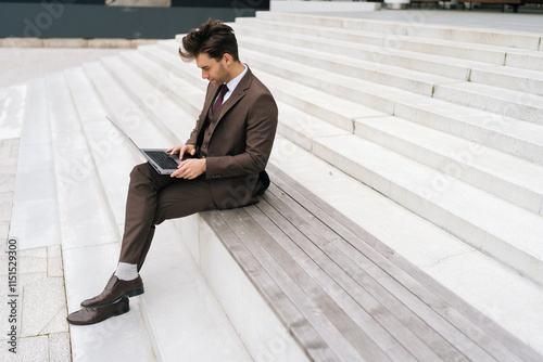 Fotografie Wide shot of focused businessman working remotely while sitting on steps in urban setting, typing on laptop computer, focused looking to screen
