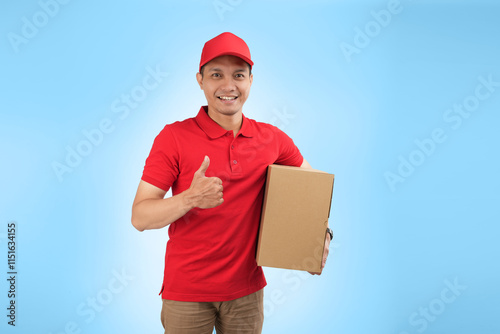 An happy Asian courier gift a thumb while holding a package against a blue background wearing red company uniform.
