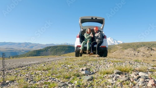 A man and a woman are relaxing, sitting in the open trunk of their car and drinking from a thermos. Tourist stop