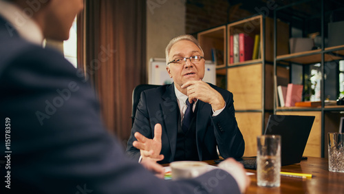 Photos Focused businessman in his late 50s sitting at table in office, engaged in lively conversation with young male employee