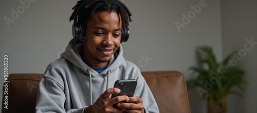 An urban cool teenager in a hoodie and dreadlocks wears headphones while engrossed in his mobile device indoors
