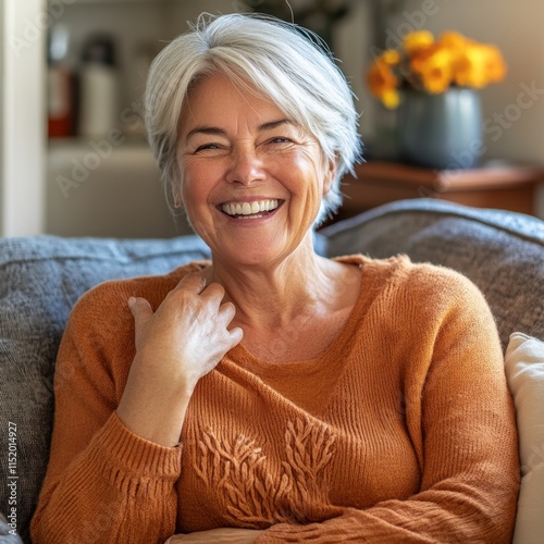 A cheerful older woman sits on her cozy home couch, smiling broadly with her perfect white teeth and laughing with a hand-on-chest gesture. photo