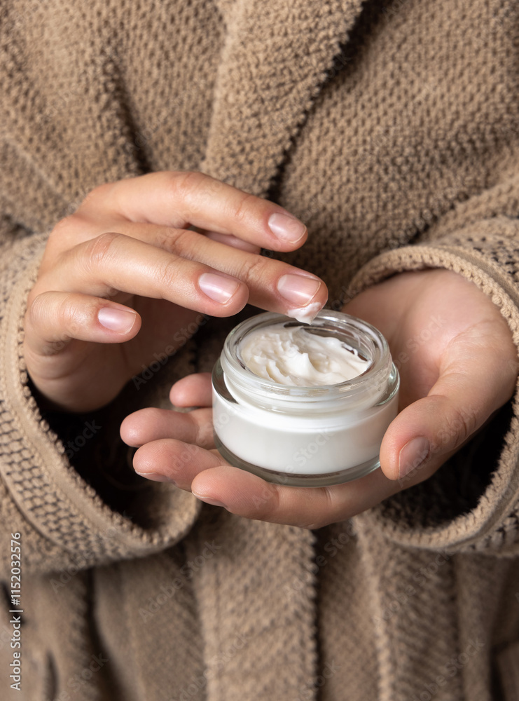 Woman in brown bathrobe touching cream with finger and holding a cosmetic jar closeup Woman in brown bathrobe touching cream with finger and holding a cosmetic jar closeup