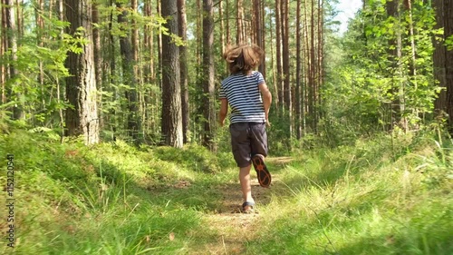 Little boy teenager with long hair in striped t-shirt, shorts and sandals running along path in coniferous forest during warm sunny summer day. Back rear following cinematic shot view