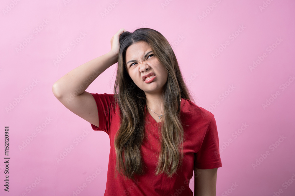 Young woman in a red shirt scratches her head with a confused or frustrated expression against a pink background. Her body language and facial expression convey uncertainty or irritation.
