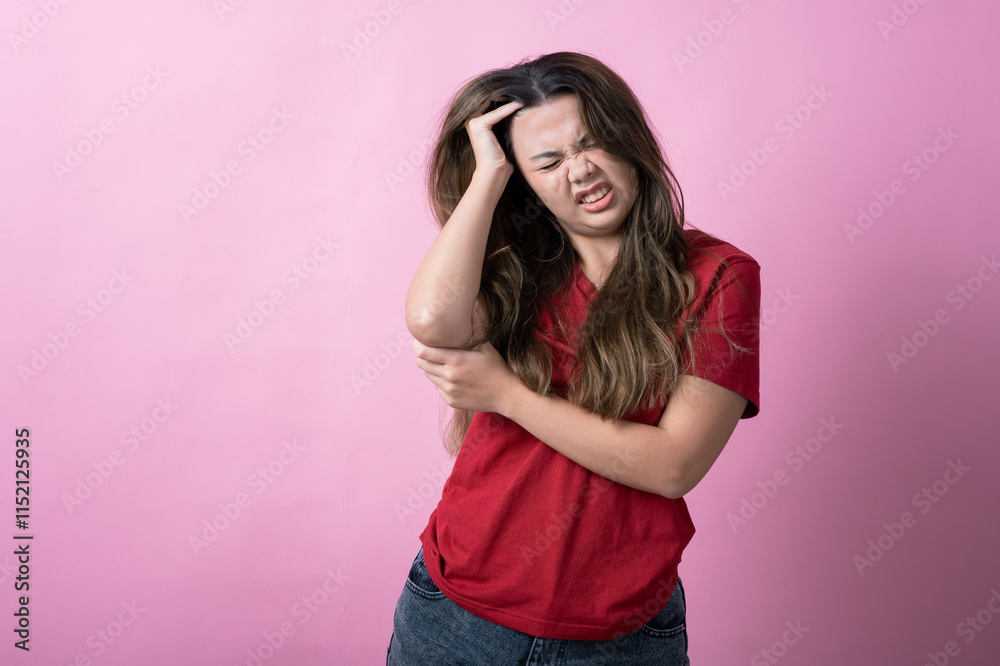 Asian woman in a red shirt with long brown hair grimaces in frustration, holding her head with one hand and grabbing her elbow with the other, standing against a solid pink background. Asian woman in a red shirt with long brown hair grimaces in frustration, holding her head with one hand and grabbing her elbow with the other, standing against a solid pink background.