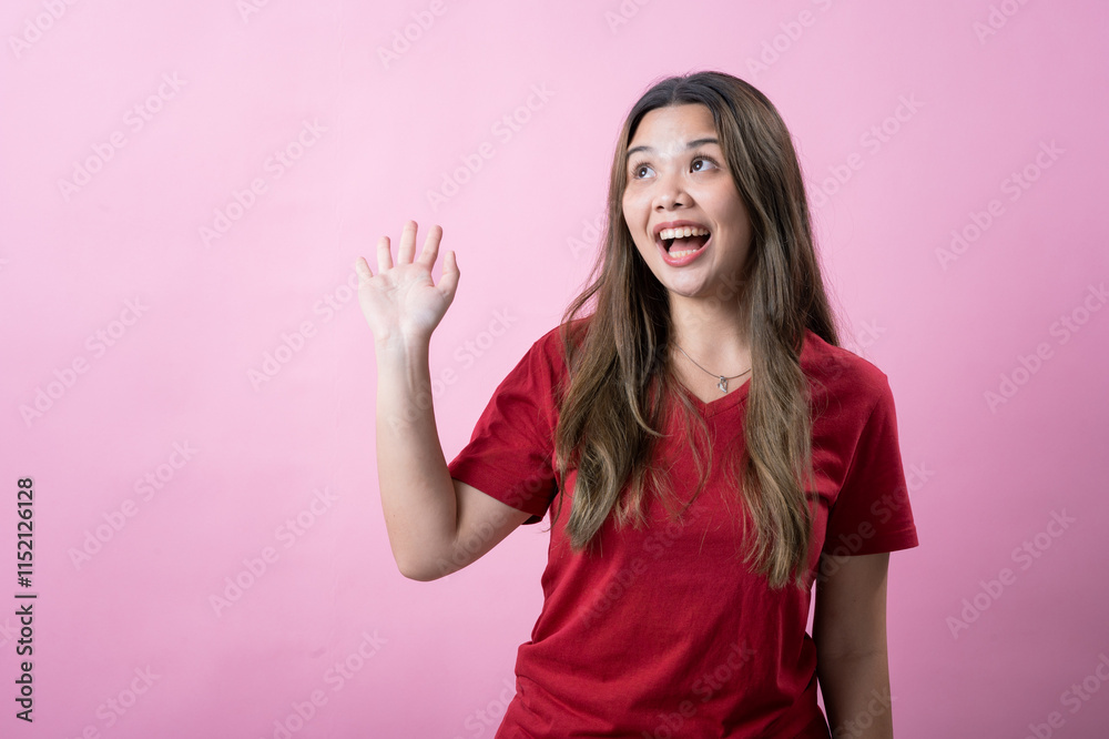 Smiling young woman with long brown hair wearing a red t-shirt waves cheerfully against a pink background. She looks excited and friendly, conveying positivity and a welcoming attitude. Smiling young woman with long brown hair wearing a red t-shirt waves cheerfully against a pink background. She looks excited and friendly, conveying positivity and a welcoming attitude.