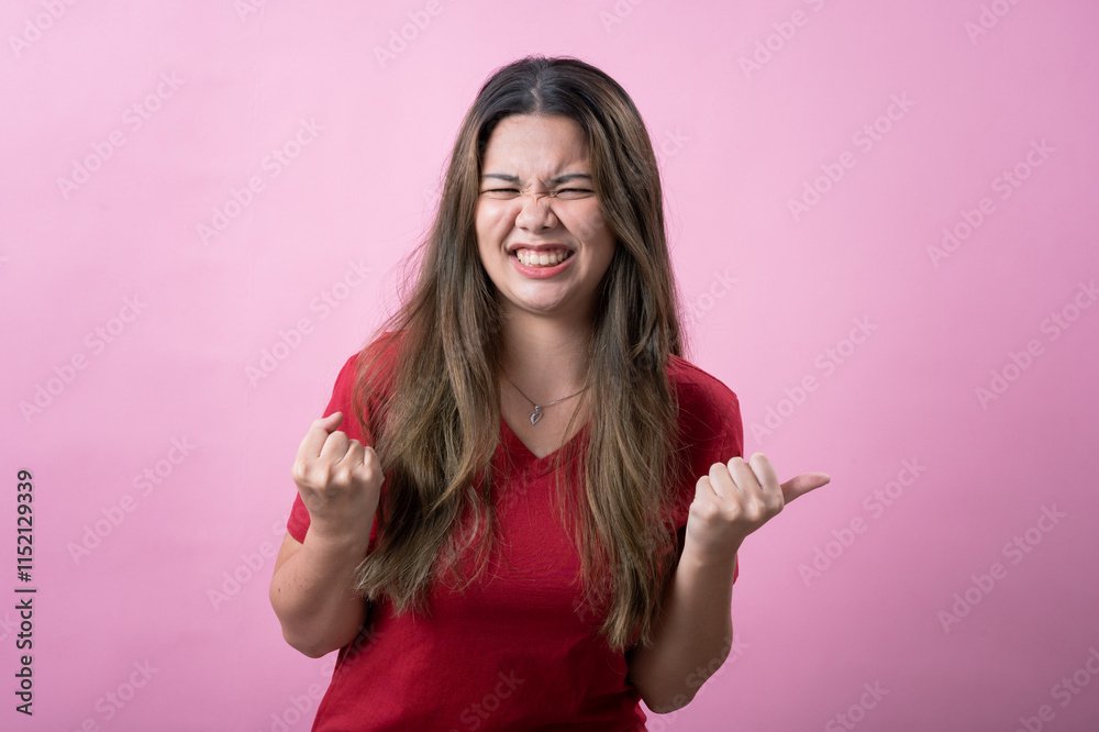 A frustrated young woman with long brown hair and light skin, wearing a red T-shirt, clenches her fists and squints her face in irritation or anger, against a solid pink background. A frustrated young woman with long brown hair and light skin, wearing a red T-shirt, clenches her fists and squints her face in irritation or anger, against a solid pink background.