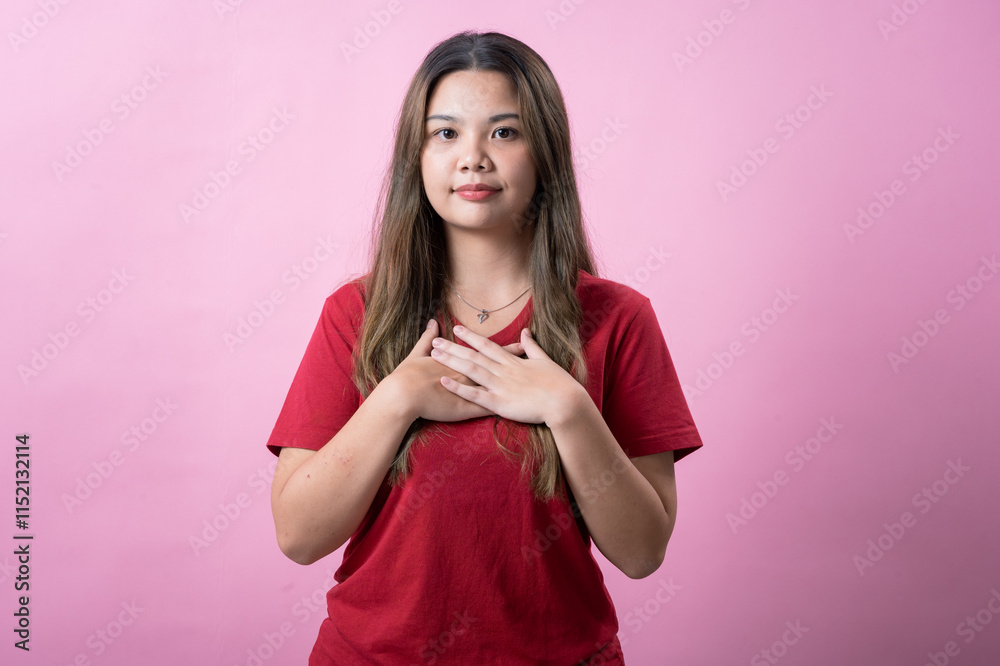 A young woman with long brown hair and light skin, wearing a red T-shirt, places her hands over her chest with a sincere and thoughtful expression, standing against a solid pink background. A young woman with long brown hair and light skin, wearing a red T-shirt, places her hands over her chest with a sincere and thoughtful expression, standing against a solid pink background.