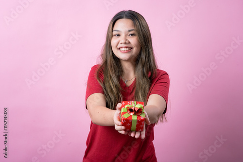 A cheerful young woman with long brown hair and light skin, wearing a red T-shirt, holds a small red gift box with a green ribbon and a decorative bow, smiling brightly against a pink background. photo