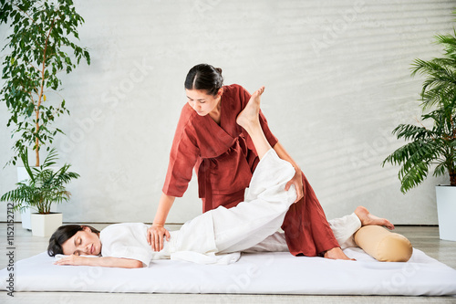 Masseuse making thai yoga massage. Woman therapist doing traditional massage treatment, stretching female patient's body. Client lying on mat, enjoying hand massage in calm atmosphere.