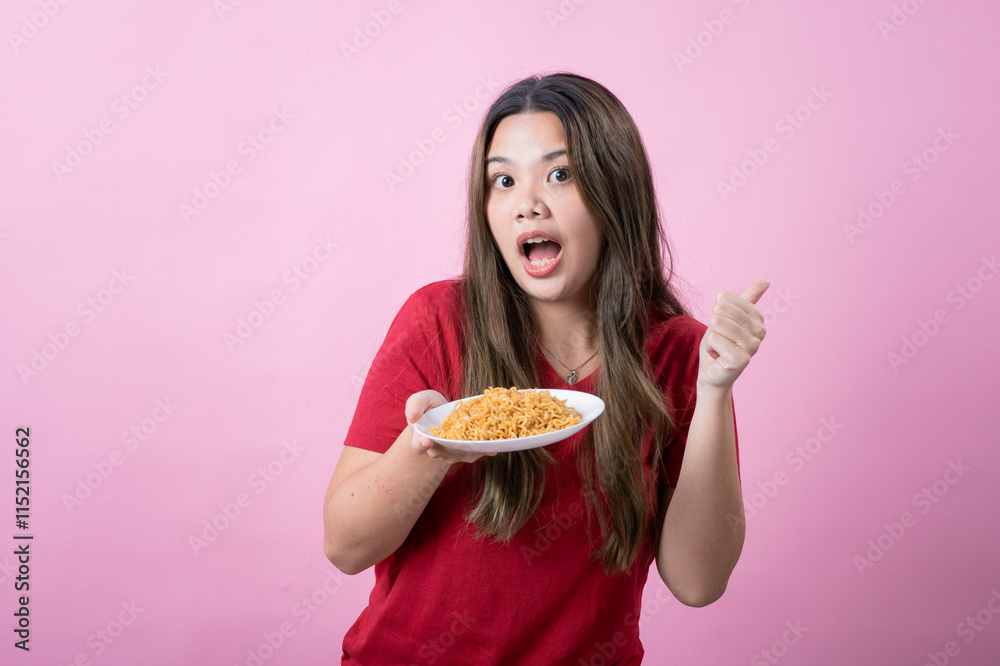 Happy young woman with long brown hair wearing a red t-shirt holding a plate of instant noodles while making an Happy young woman with long brown hair wearing a red t-shirt holding a plate of instant noodles while making an