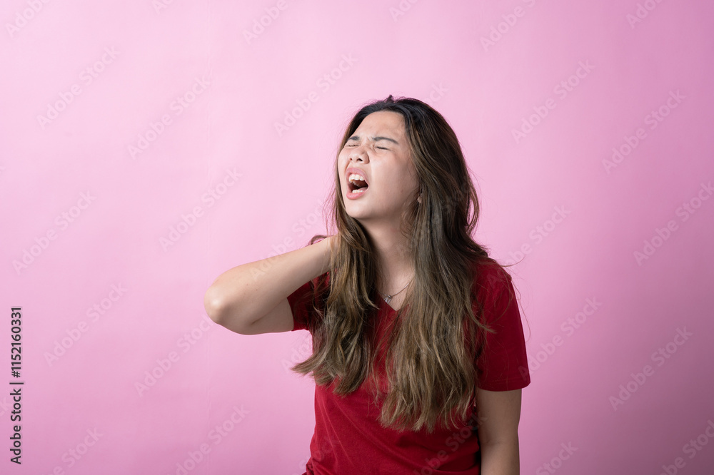 Asian woman in a red shirt with long brown hair grimaces in pain, holding the back of her neck with one hand, against a solid pink background. Asian woman in a red shirt with long brown hair grimaces in pain, holding the back of her neck with one hand, against a solid pink background.