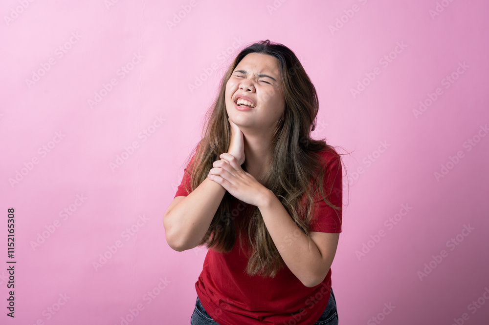 Asian woman in a red shirt with long brown hair grimaces in pain, holding the back of her neck with one hand, against a solid pink background. Asian woman in a red shirt with long brown hair grimaces in pain, holding the back of her neck with one hand, against a solid pink background.