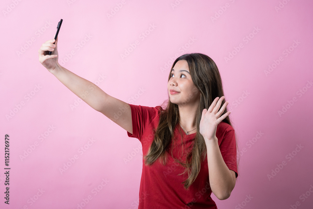 Excited young woman with long brown hair in a red t-shirt waves while taking a selfie with a smartphone against a pink background. She looks cheerful and friendly, capturing a fun and lively moment. Excited young woman with long brown hair in a red t-shirt waves while taking a selfie with a smartphone against a pink background. She looks cheerful and friendly, capturing a fun and lively moment.