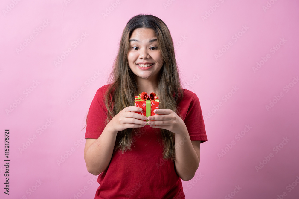 A cheerful young woman with long brown hair and light skin, wearing a red T-shirt, holds a small red gift box with a green ribbon and a decorative bow, smiling brightly against a pink background. A cheerful young woman with long brown hair and light skin, wearing a red T-shirt, holds a small red gift box with a green ribbon and a decorative bow, smiling brightly against a pink background.
