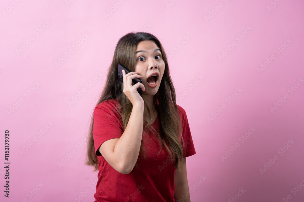 Surprised young woman with long brown hair wearing a red t-shirt talks on a smartphone against a pink background, reacting with shock or excitement during a phone call, expressing strong emotions. Surprised young woman with long brown hair wearing a red t-shirt talks on a smartphone against a pink background, reacting with shock or excitement during a phone call, expressing strong emotions.