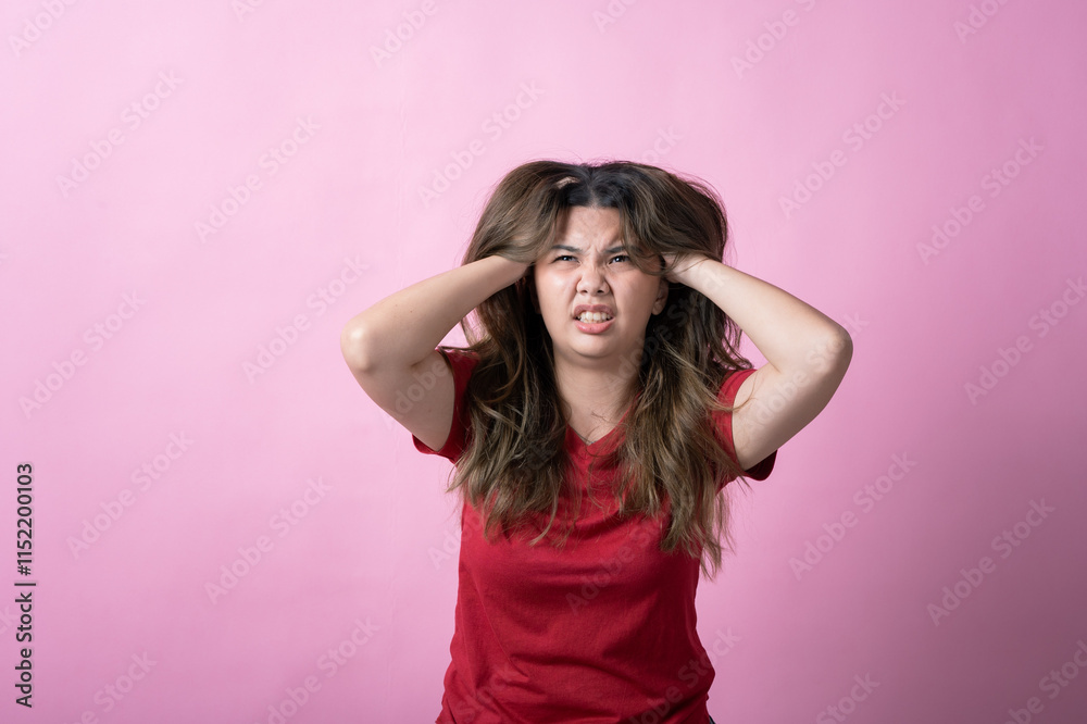 Asian woman in a red shirt with long brown hair grips her head with both hands, eyes closed, and mouth open in frustration against a pink background. Asian woman in a red shirt with long brown hair grips her head with both hands, eyes closed, and mouth open in frustration against a pink background.
