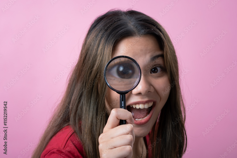 A young woman with long brown hair , wearing a red T-shirt, smiles playfully while holding a magnifying glass up to her eye, enlarging it for a fun and curious effect, against a pink background. A young woman with long brown hair , wearing a red T-shirt, smiles playfully while holding a magnifying glass up to her eye, enlarging it for a fun and curious effect, against a pink background.